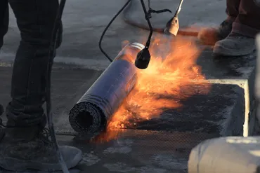 Person using a torch to apply heat on rolled roofing material with flames visible on a roof surface.