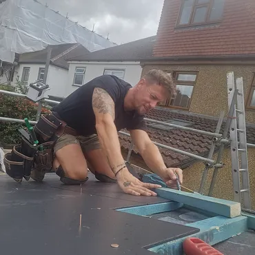 Man in shorts and black shirt working on roof with tools and scaffolding nearby on a cloudy day.