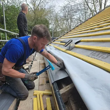 A man in a blue shirt uses a hammer to secure roofing material on a sloped roof under construction.