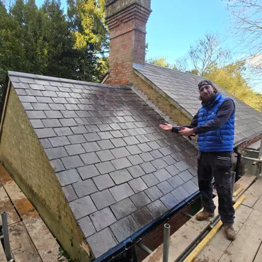Man in blue vest and sunglasses standing on scaffolding next to newly cleaned slate roof with chimney.