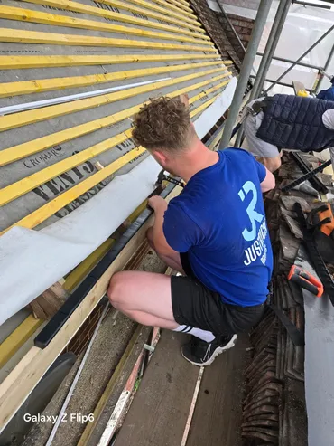 Man in blue shirt kneeling and working on wooden strips on a roof under construction.