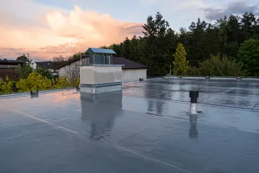 Flat roof with reflective waterproof membrane and ventilation pipes at sunset with trees in background.