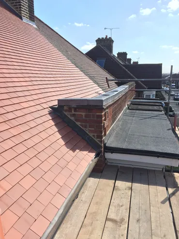 Roof with red tiles, brick chimney, flat black roof section, and wooden scaffolding floor under sunlight.