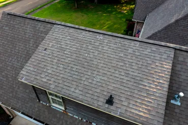 Aerial view of a gray shingled roof of a residential house with a street and lawn visible above.
