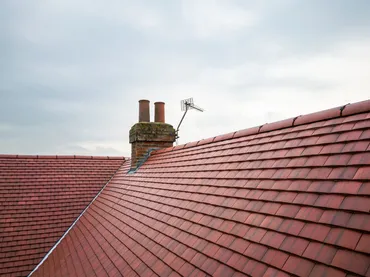 View of reddish roof tiles with a brick chimney and a TV antenna under a cloudy sky.