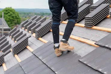 Person wearing boots and pants installing dark roof tiles on a sloped roof outdoors.