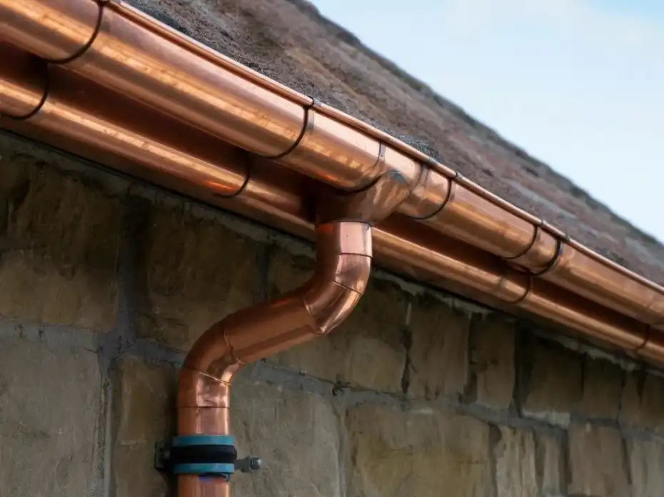 Copper gutter system attached to stone wall under roof edge with blue sky background.