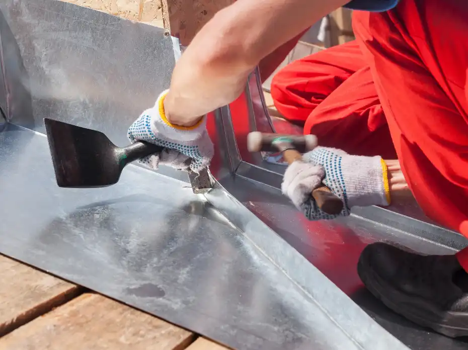 Person wearing gloves and red pants working with hammer and tool on sheet metal installation.