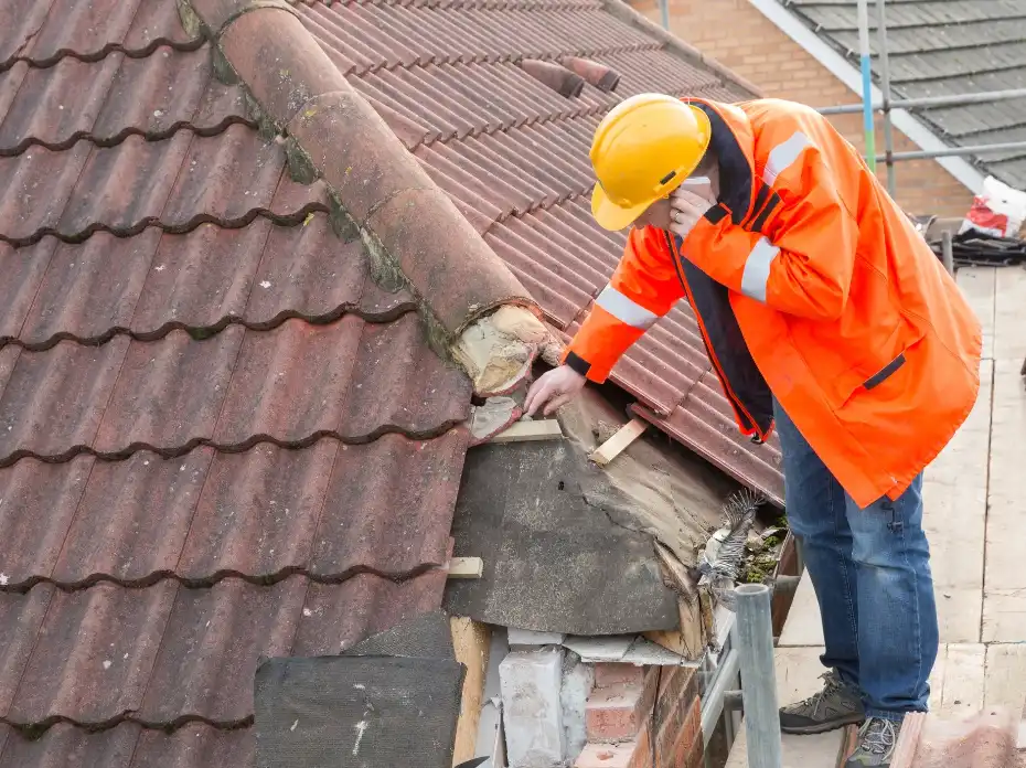 Worker in orange jacket and yellow helmet inspecting damaged roof tiles on a house roof.